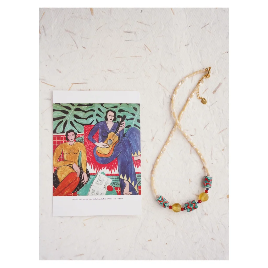 A beaded necklace with green ceramic beads and yellow glass next to a Matisse print.