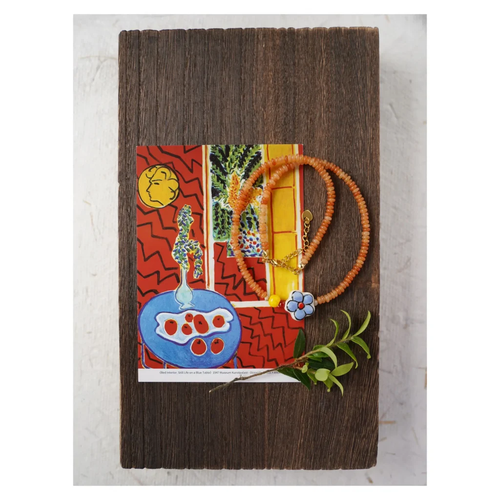 Matisse Series: Handmade red aventurine necklace with a blue floral ceramic bead, displayed on a vintage wood block with Henri Matisse's Red Interior artwork, by Jewea.