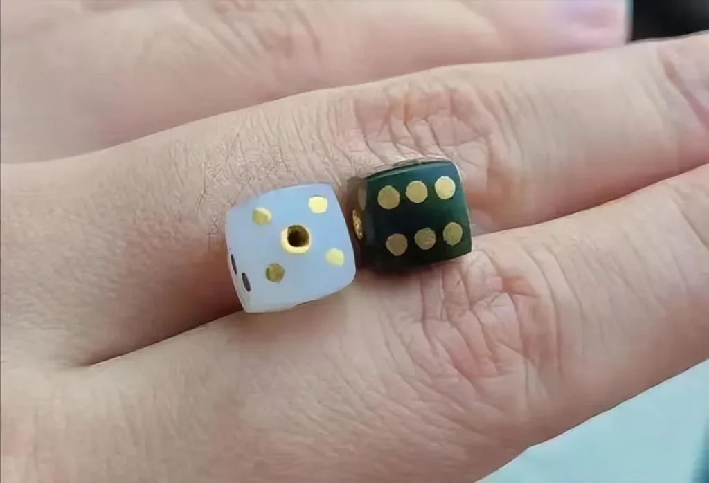 An illustration of a dice pattern, specifically showing two small, square dice resting on a finger. One die is light-colored (off-white or light jade) with gold-colored dots, and the other is dark green with gold-colored dots, reflecting the look of carved, decorative dice.