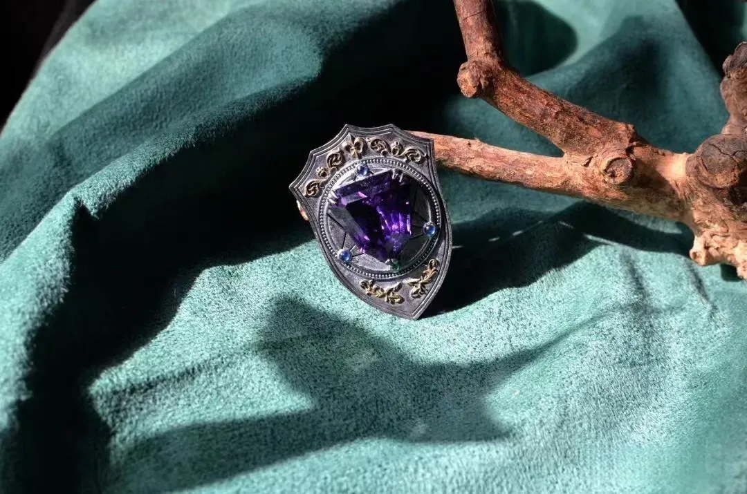 A vintage-style gothic ring featuring a large, shield-cut purple amethyst gemstone set in an oxidized silver frame with intricate gold filigree details.