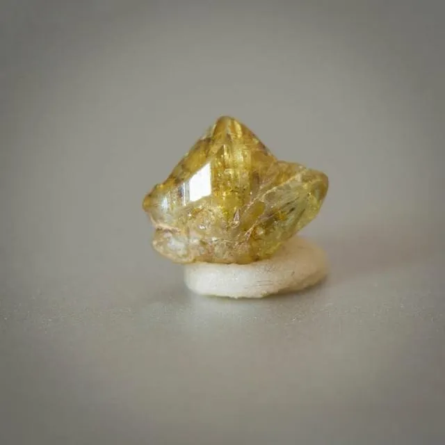 An image of a chrysoberyl knee-shaped twin crystal specimen. The crystal has a bent shape resembling a knee, which is a common twinning habit for chrysoberyl, and its color is typically yellowish-green or brownish-yellow.