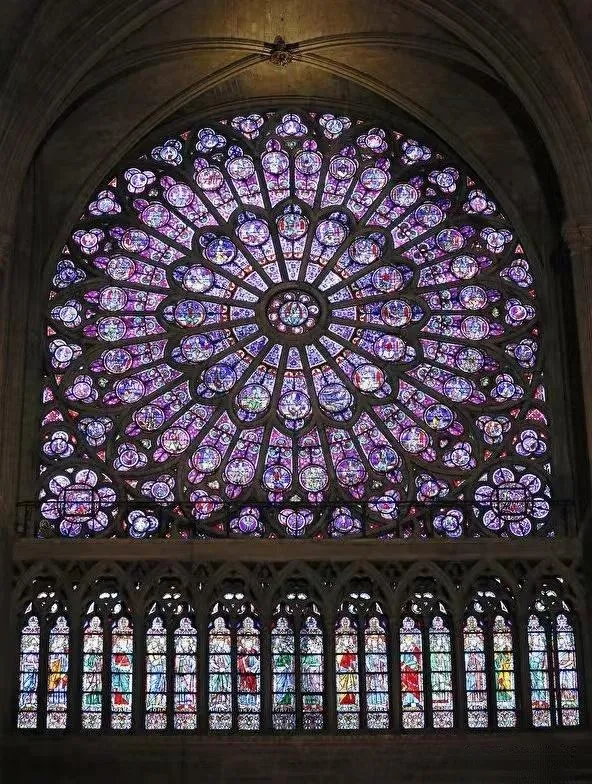 An illustration of the rose window of Notre-Dame Cathedral. The window is characterized by its massive and ornate circular design, composed of countless pieces of stained glass depicting biblical stories and figures of saints. Light shines through the glass, casting brilliant and colorful patterns inside the cathedral.
