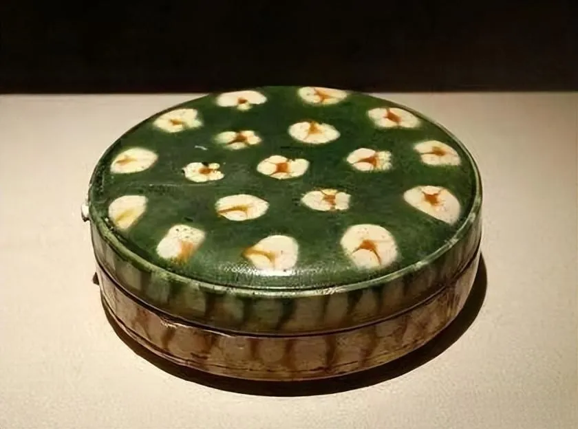 An illustration of a Tang Sancai powder box. The small, round ceramic box is decorated with the signature vibrant glazes in shades of green, amber, and cream, showcasing the iconic artistry of the Tang Dynasty.