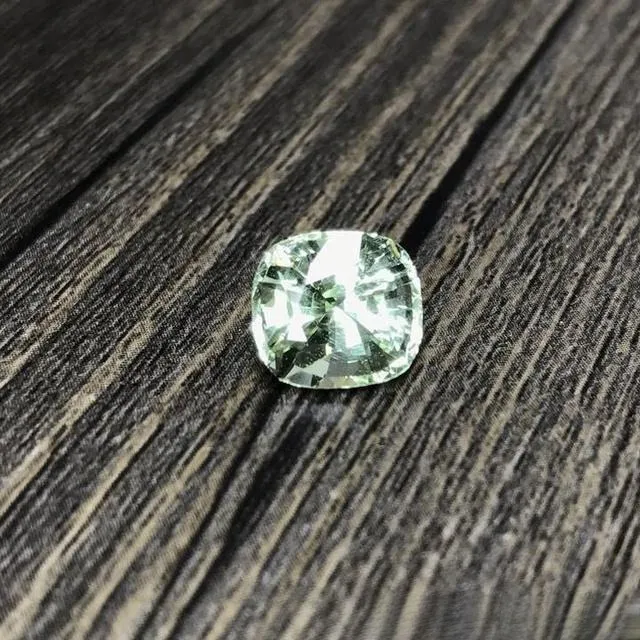 A real photo of a loose vanadium chrysoberyl gemstone placed on a solid wood tabletop. The gem displays a vibrant green color, similar to a green apple, which contrasts with the warm texture of the wooden table, highlighting the gemstone's rarity and natural beauty.