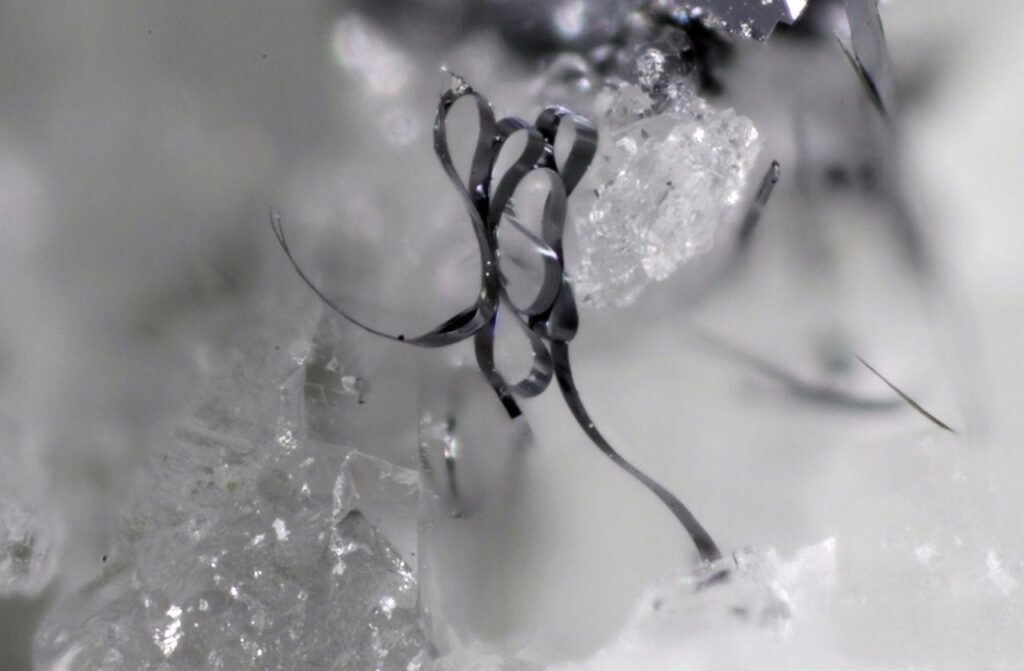 Microscopic view of bismuth inclusions curled like tape inside quartz, with a smooth metal-like surface impossible to replicate with real steel