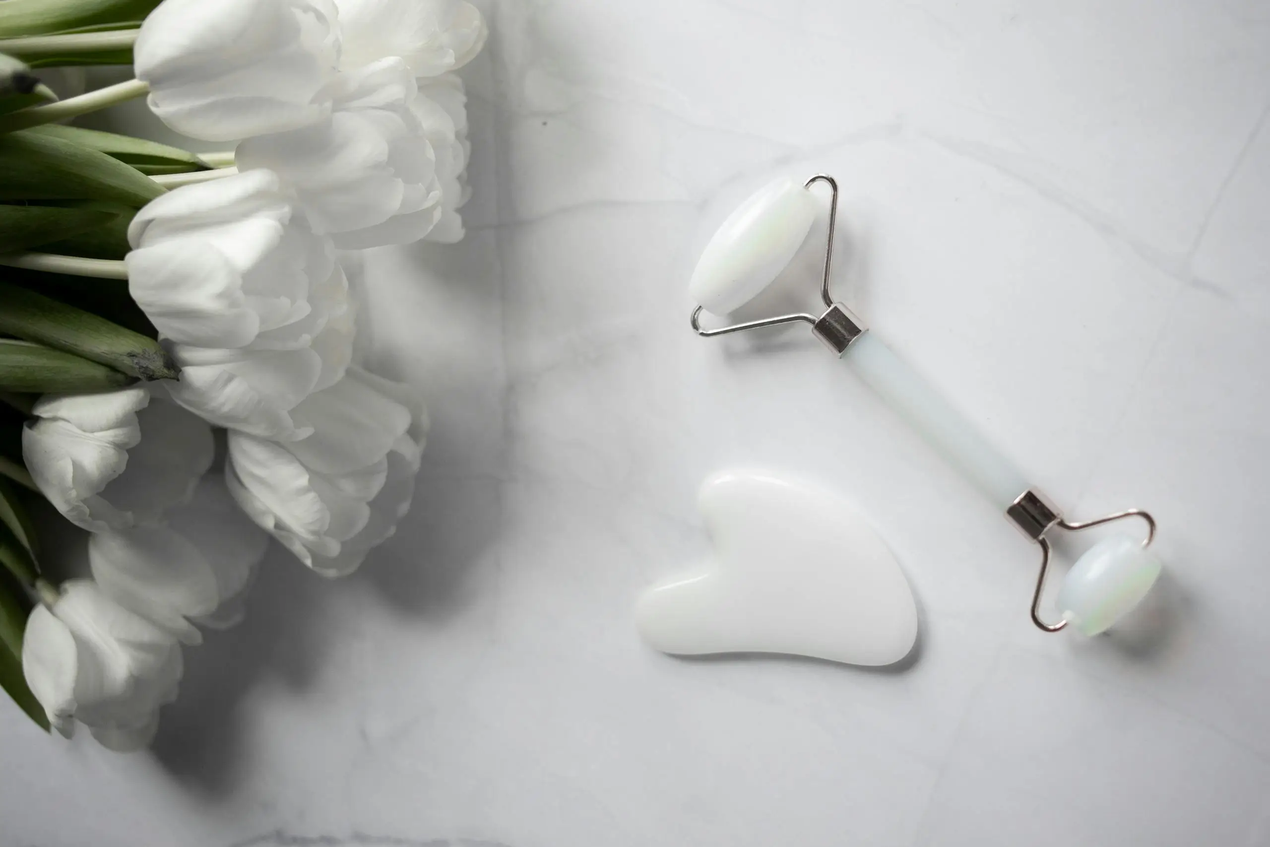White tulips with jade roller and gua sha on marble, illustrating the natural beauty of jade in skincare.