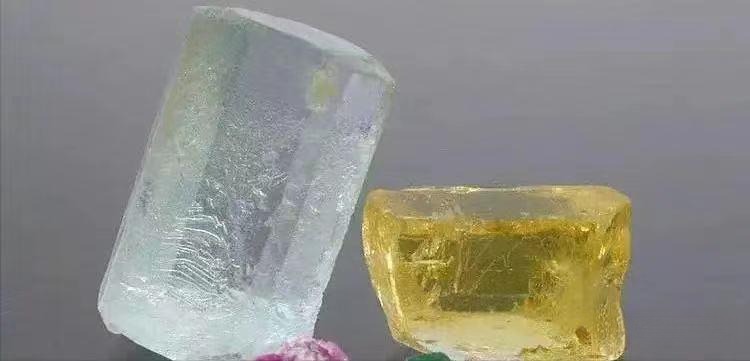 Two large Beryl rough stones: a blue Brazilian Aquamarine on the left and a yellow Heliodor, showcasing their natural crystal forms.