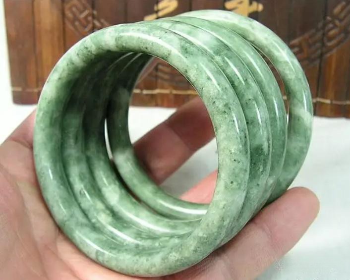 A person's hand holding four low-grade Dushan Jade bangles, notable for their opaque quality and mottled, inconsistent coloring.