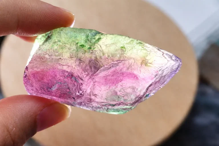 A hand holding a multi-colored rainbow fluorite crystal showing purple and green banding.