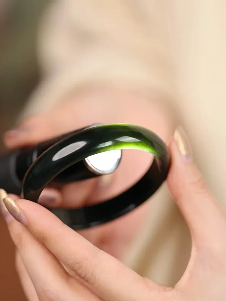 A dark Hei Qing nephrite bracelet showing greenish-yellow light transmission under backlighting.