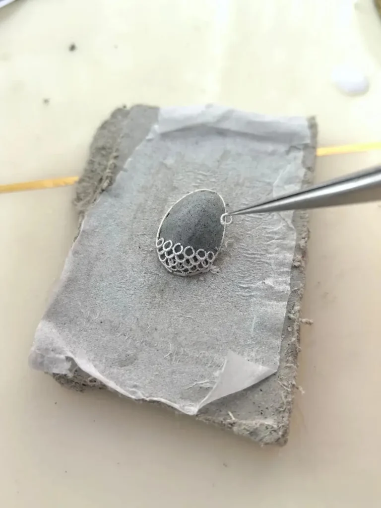 An artisan using tweezers to stack gold filigree layers over a clay base for 3D shaping.