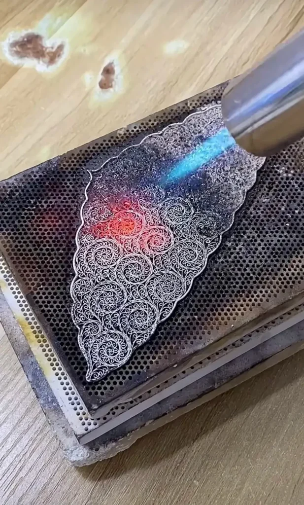 An artisan using a blowtorch to fuse microscopic solder joints on a gold filigree frame.