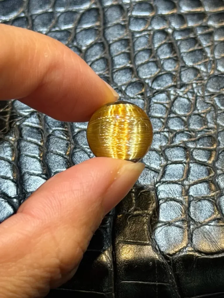 A 14.4mm Titanium Quartz bead held in fingers, showcasing metallic rutile blades and intense light reflection.