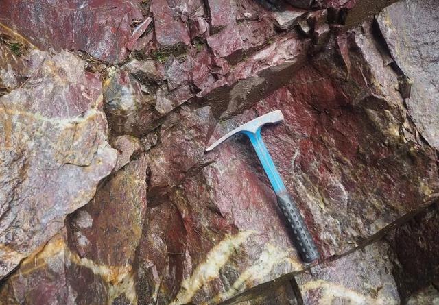 A geological hammer rests against a rugged primary deposit of raw blood-red jasper.