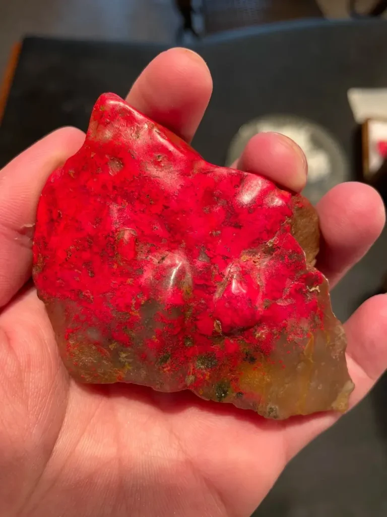 A hand holding a raw Zhejiang Changhua chicken-blood stone with vibrant red patches on a gray matrix.