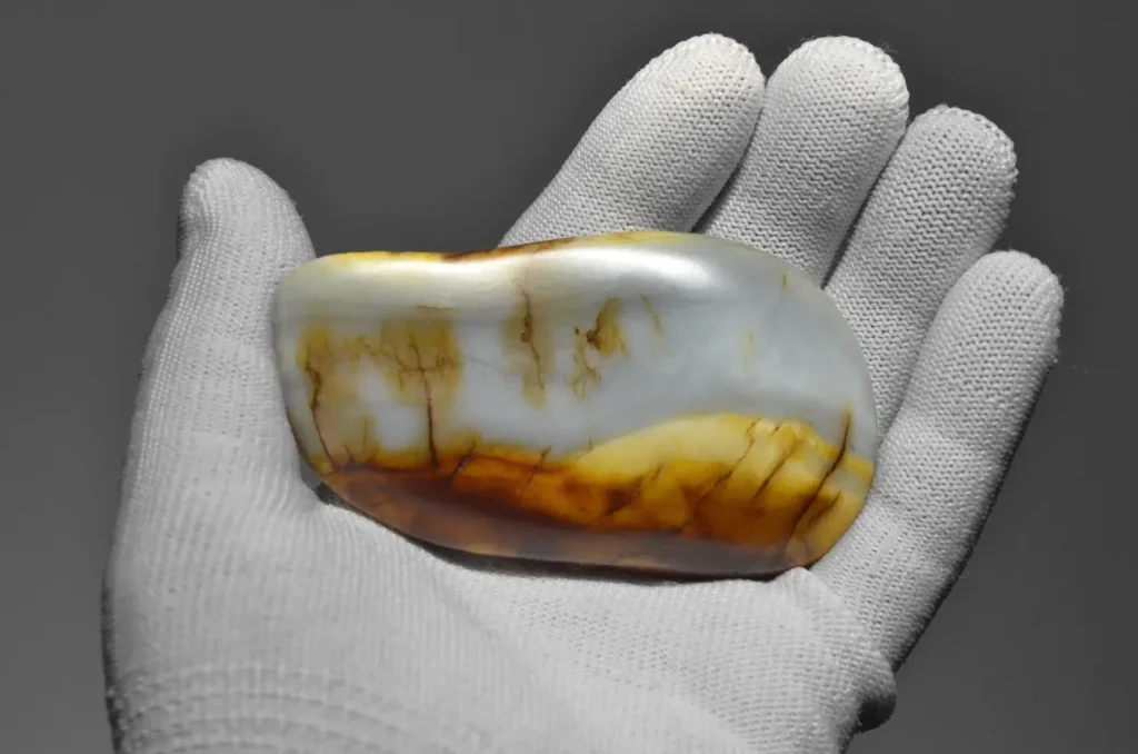 A gloved hand holds a smooth Dahua Nephrite seed stone with white and golden-brown landscape patterns.