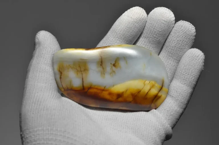 A gloved hand holds a smooth Guangxi Nephrite seed stone with white and golden-brown landscape patterns.