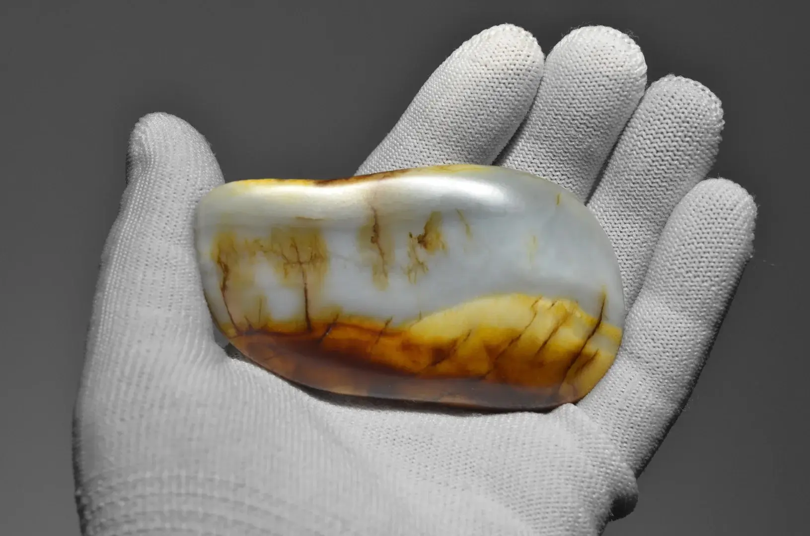A gloved hand holds a smooth Guangxi Nephrite seed stone with white and golden-brown landscape patterns.