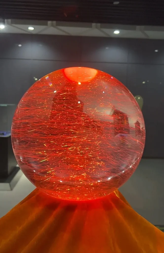 A large rutilated quartz sphere with golden-red needle inclusions, illuminated on a museum stand.