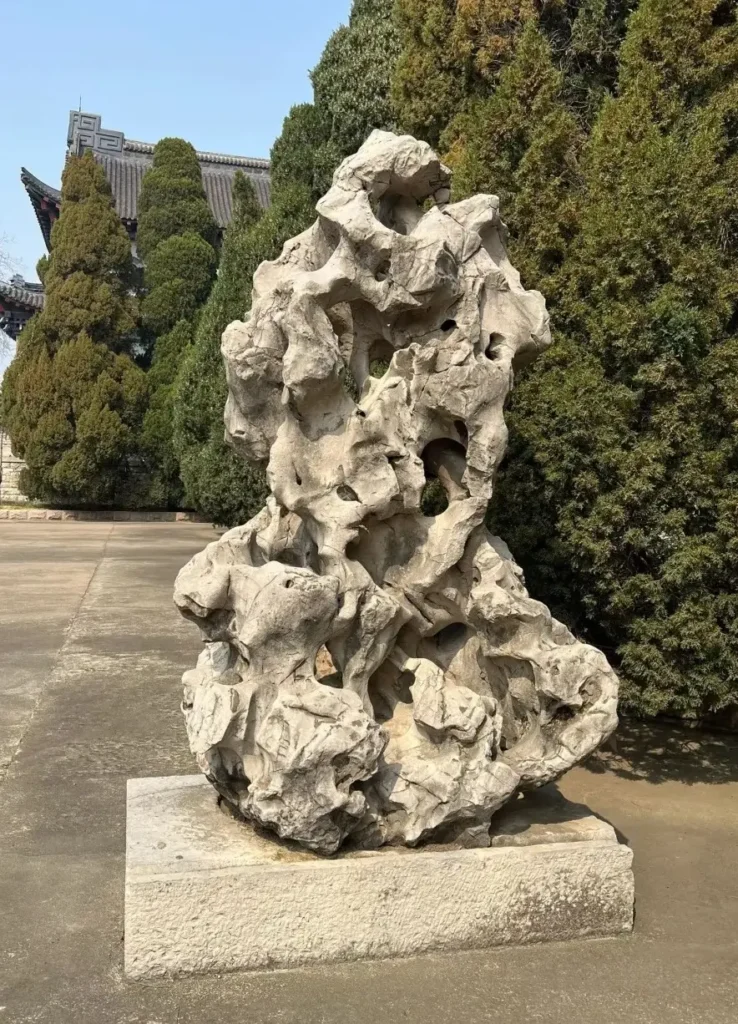 A vertical, weather-worn Jiangsu Taihu stone with complex apertures on a square stone base outdoors.