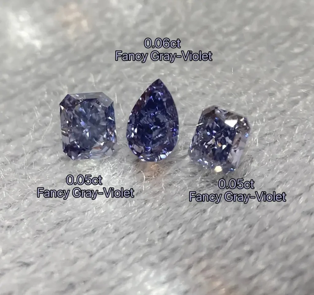 Three fancy gray-violet diamonds in radiant and pear cuts resting on a white textured surface.
