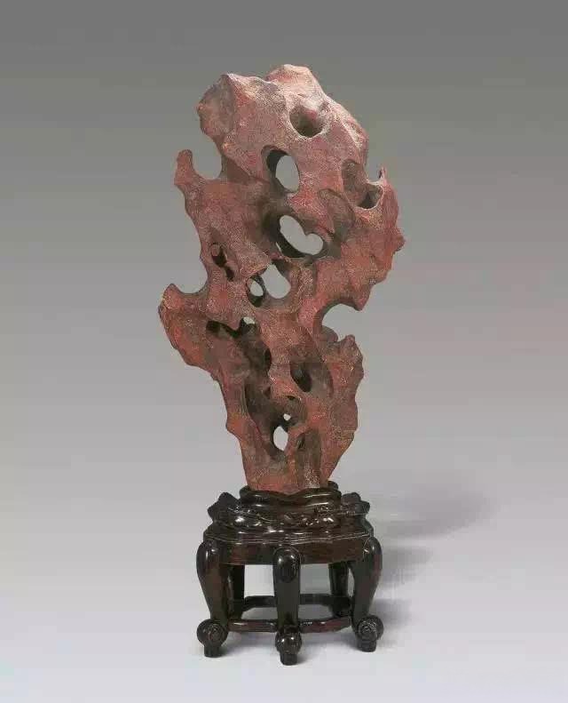 A vertical red Jiangsu Taihu stone with natural perforations, displayed on a carved dark wooden pedestal.