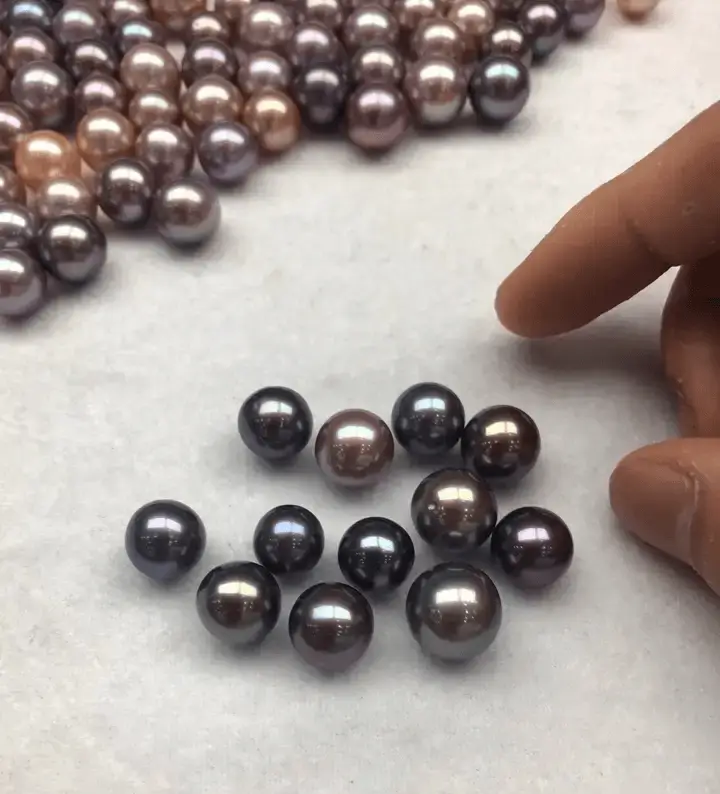A hand sorting large, high-luster freshwater gems on a white surface with a pile in the background.