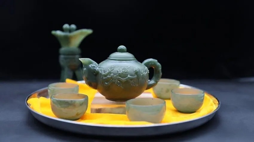 Carved grayish-green Xianghua jade teapot and teacups from Hunan, displayed on yellow cloth.