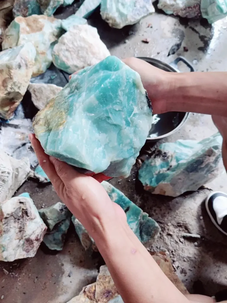 A pair of hands holding a large, raw piece of light aquamarine-blue Guicui quartzite.