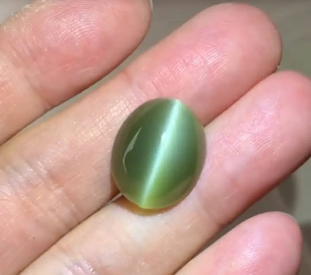 An oval green Cat’s Eye Nephrite cabochon from Taiwan held between fingers, showing a sharp chatoyant band.