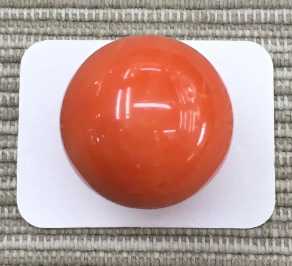 A polished 17mm round orange-red Taiwan Momo coral bead weighing 6.79 grams on a white card.
