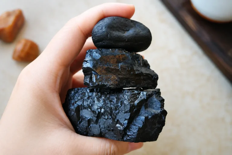 A hand holding three stacked jet stone specimens, showing both smooth tumbled and jagged raw textures.