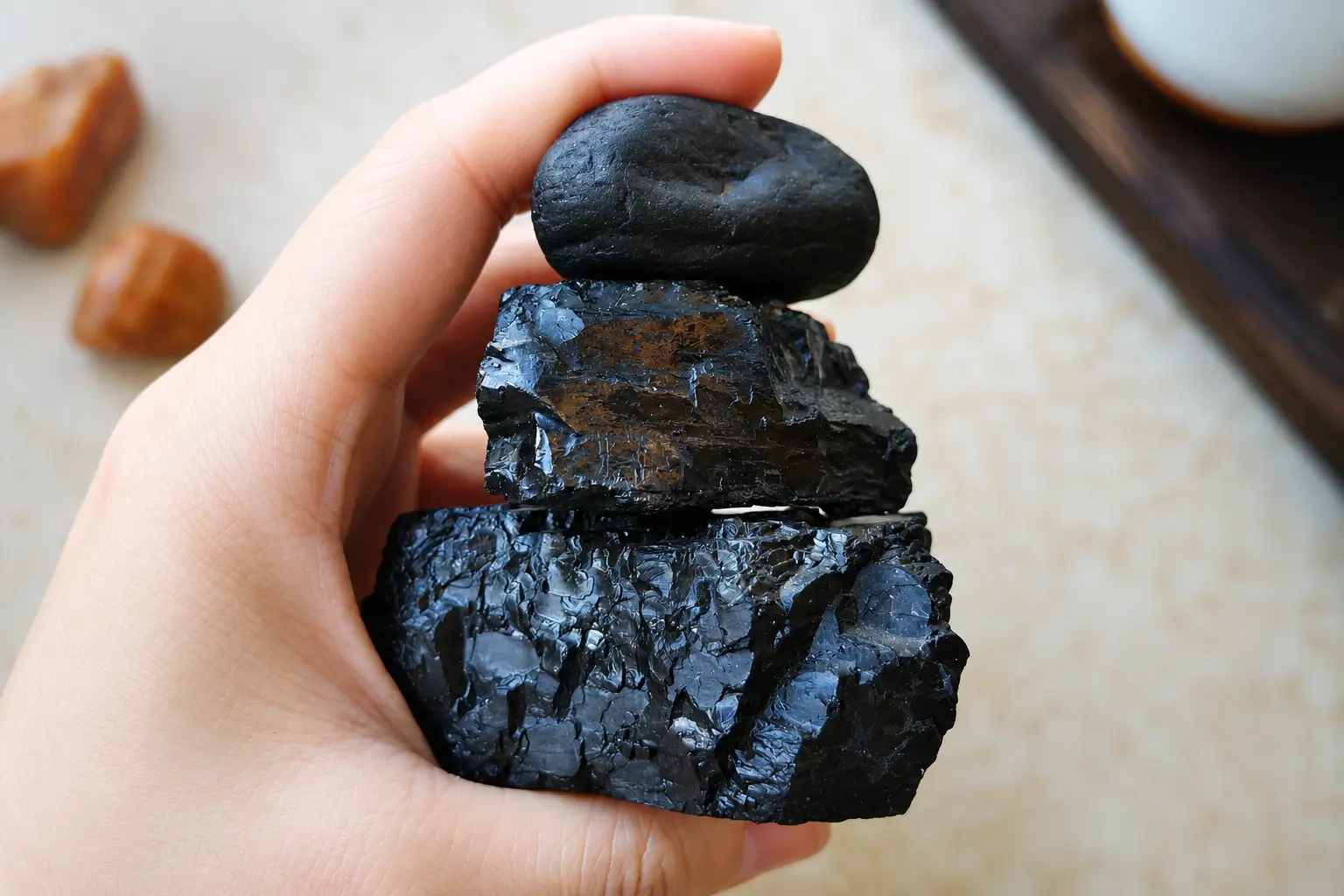 A hand holding three stacked jet stone specimens, showing both smooth tumbled and jagged raw textures.
