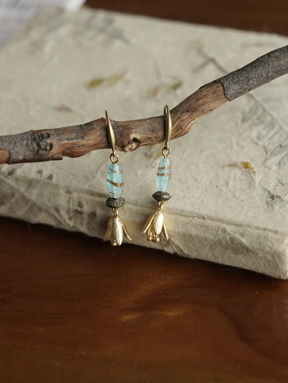 Vintage-style earrings hanging on a branch over a textured notebook on a dark wooden surface.