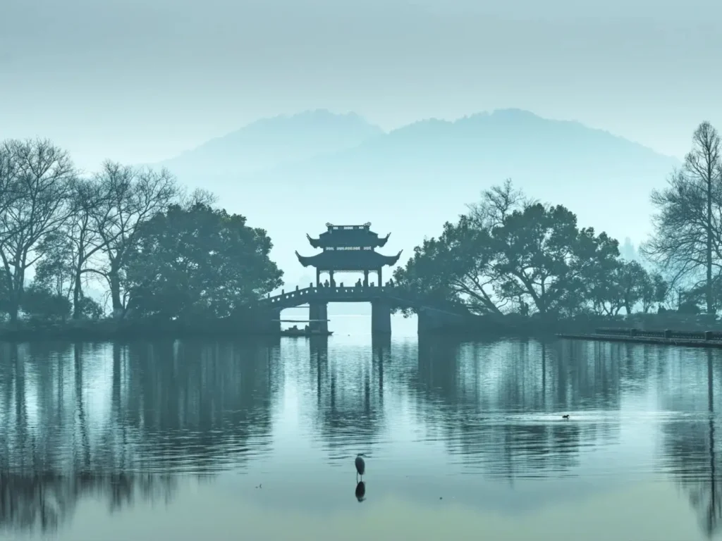 Mist covers West Lake in Hangzhou, Zhejiang, with a traditional stone arch bridge, trees, and an ornate pavilion.