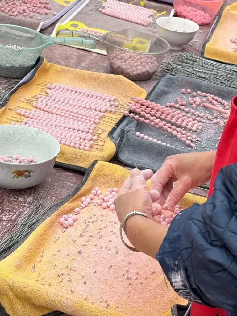 An artisan shaping pink clay beads and piercing them with a fine needle on a workshop table.