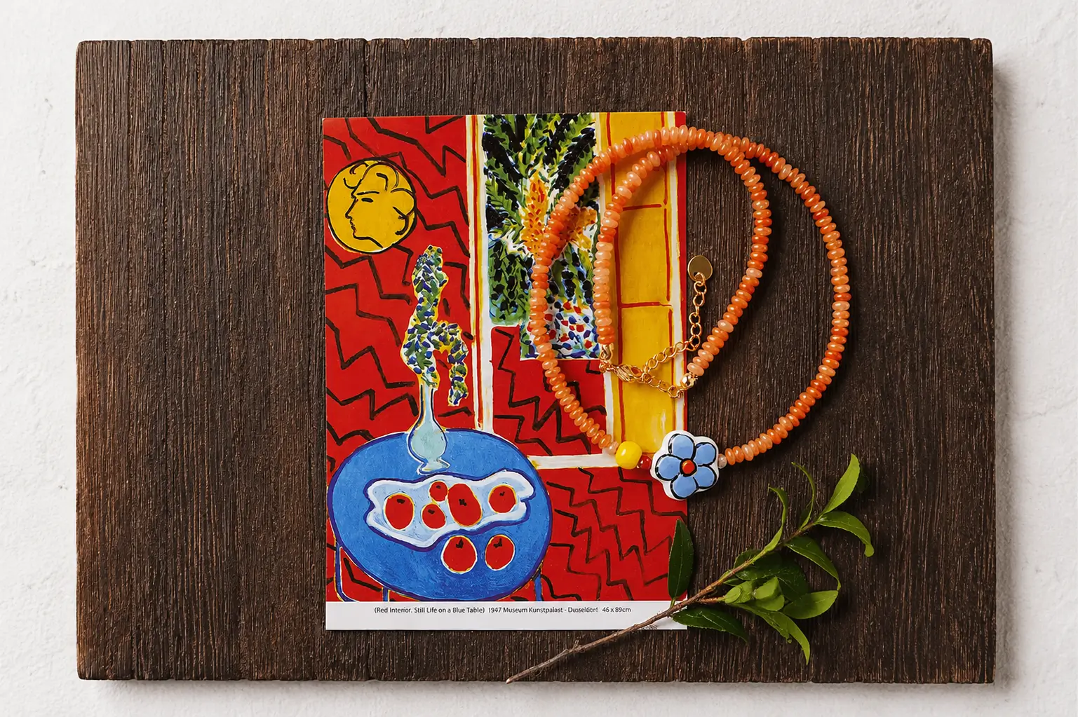 Red aventurine bead necklace on a Matisse print with a ceramic flower pendant and green sprig on dark wood.