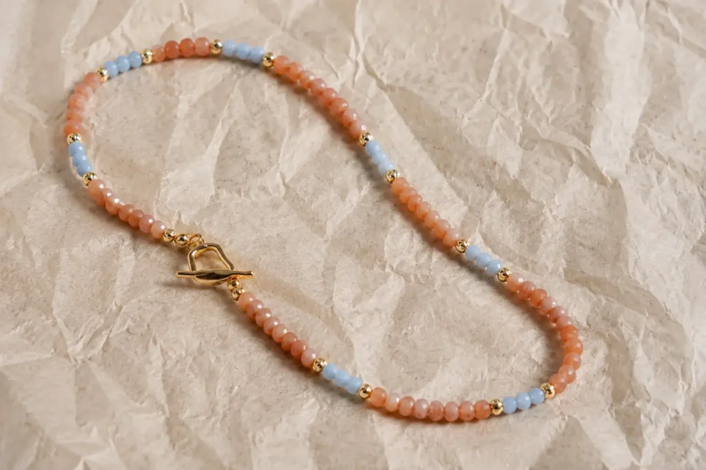 A beaded necklace with peach-orange sunstone, light blue beads, and gold beads on textured paper.