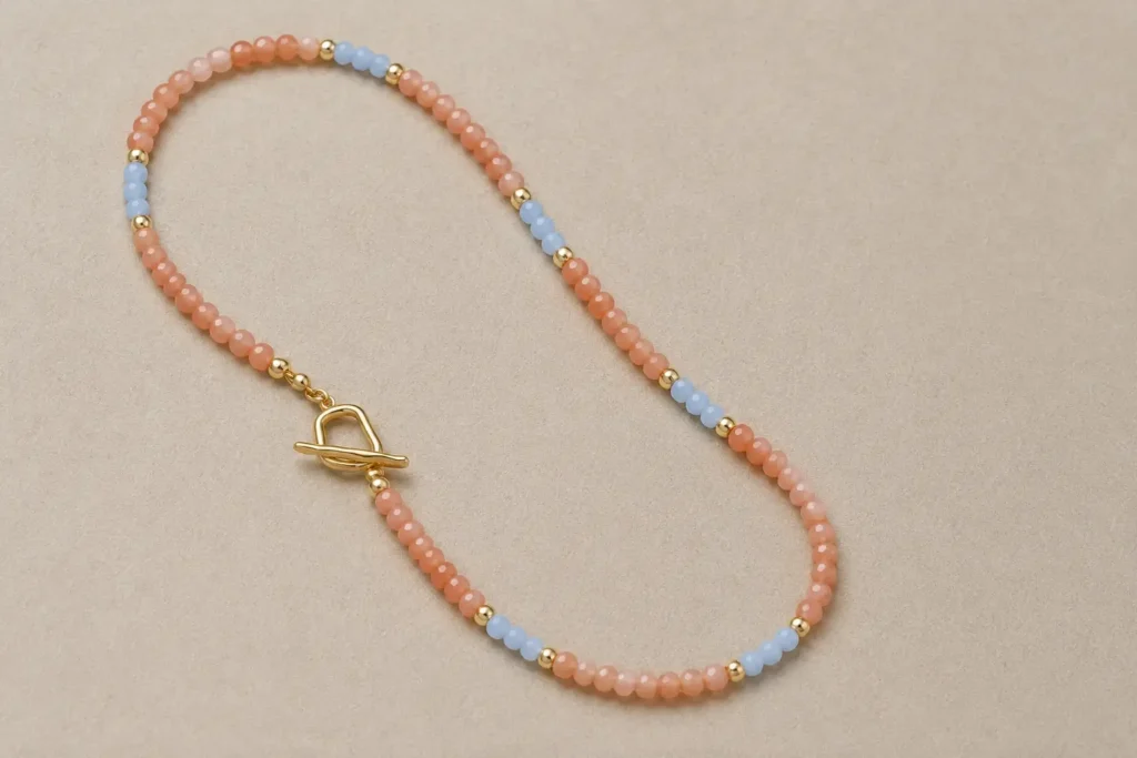 A beaded necklace with peach sunstone, light blue accents, and a gold clasp on a gray background.