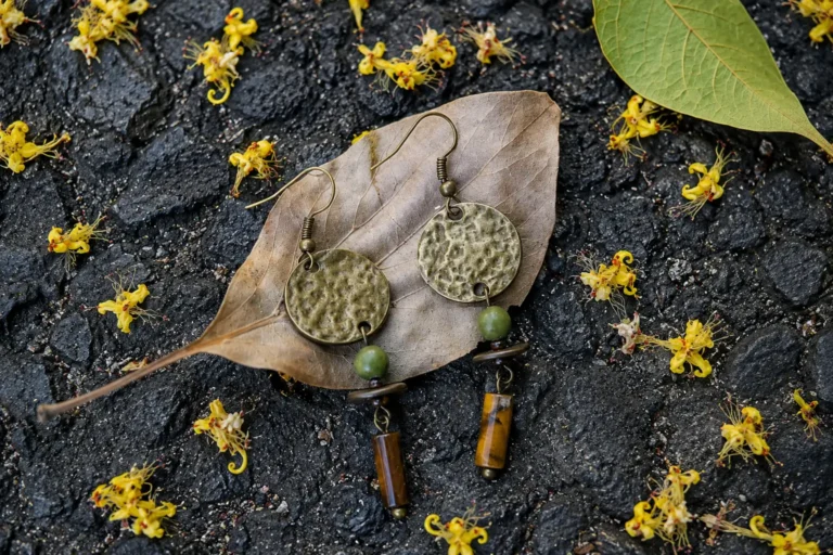 Pair of round hammered brass earrings with green and brown gemstone beads resting on a dry leaf.