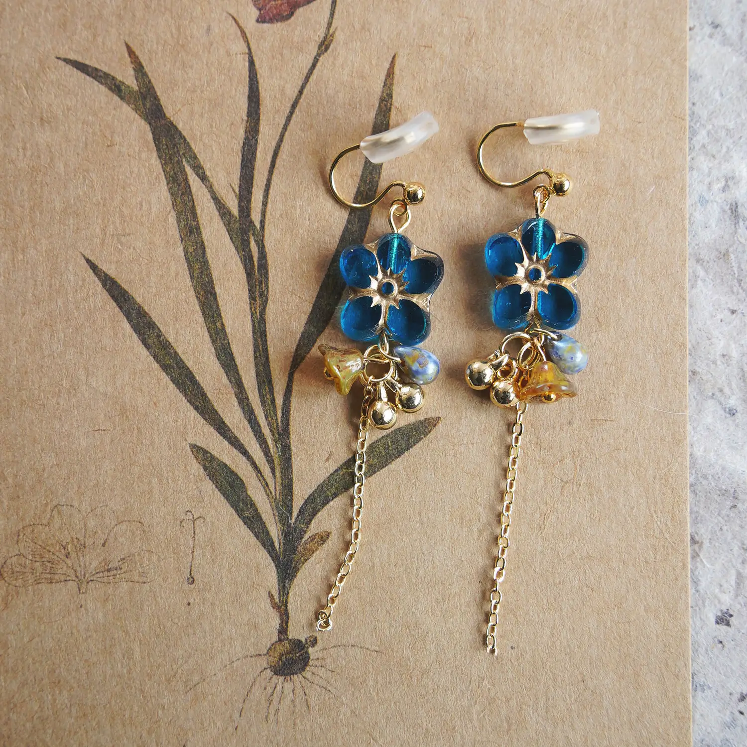 Blue glass clip-on earrings with long gold chains displayed on a botanical card.