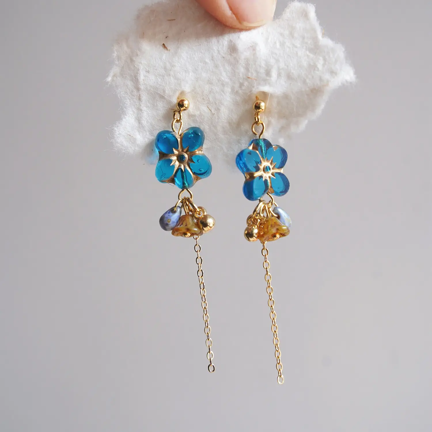 Blue glass drop earrings held above white fabric, showing handmade details and a lightweight design.