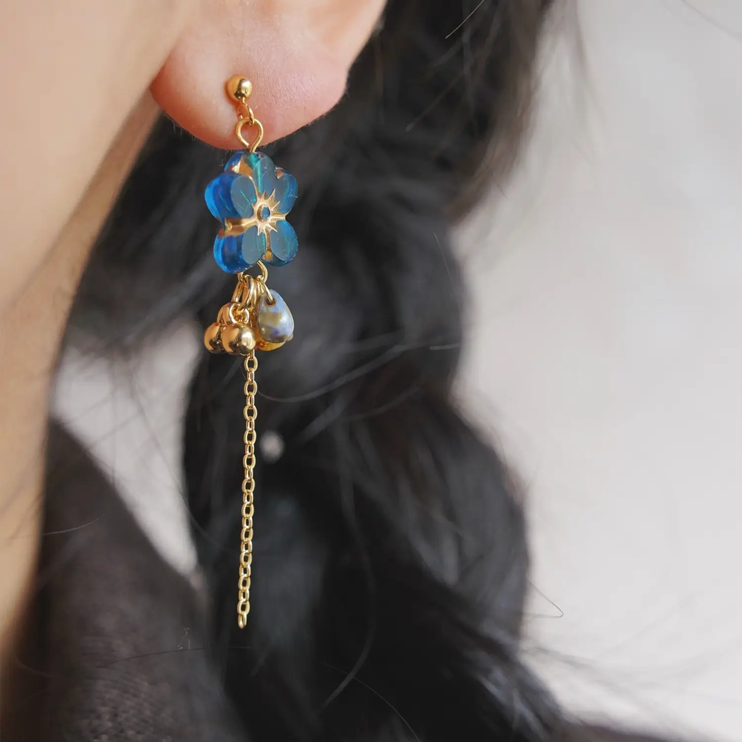 Model wearing blue glass flower drop earring with long gold chain against dark hair.