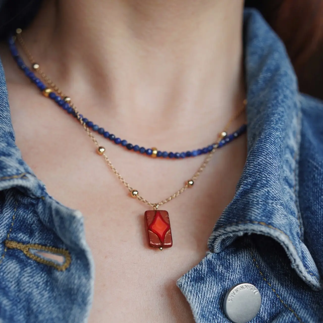 Blue beaded necklace layered with a gold pendant over a denim jacket.
