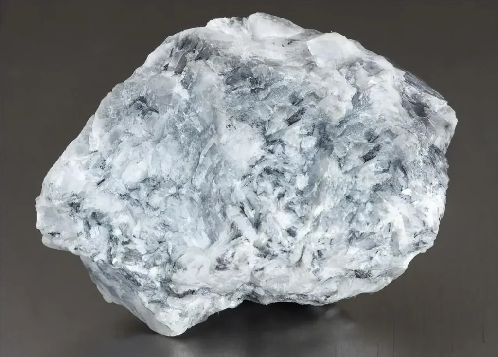 A raw light gray magnesite mineral specimen with a rough crystalline texture on a dark grey surface.
