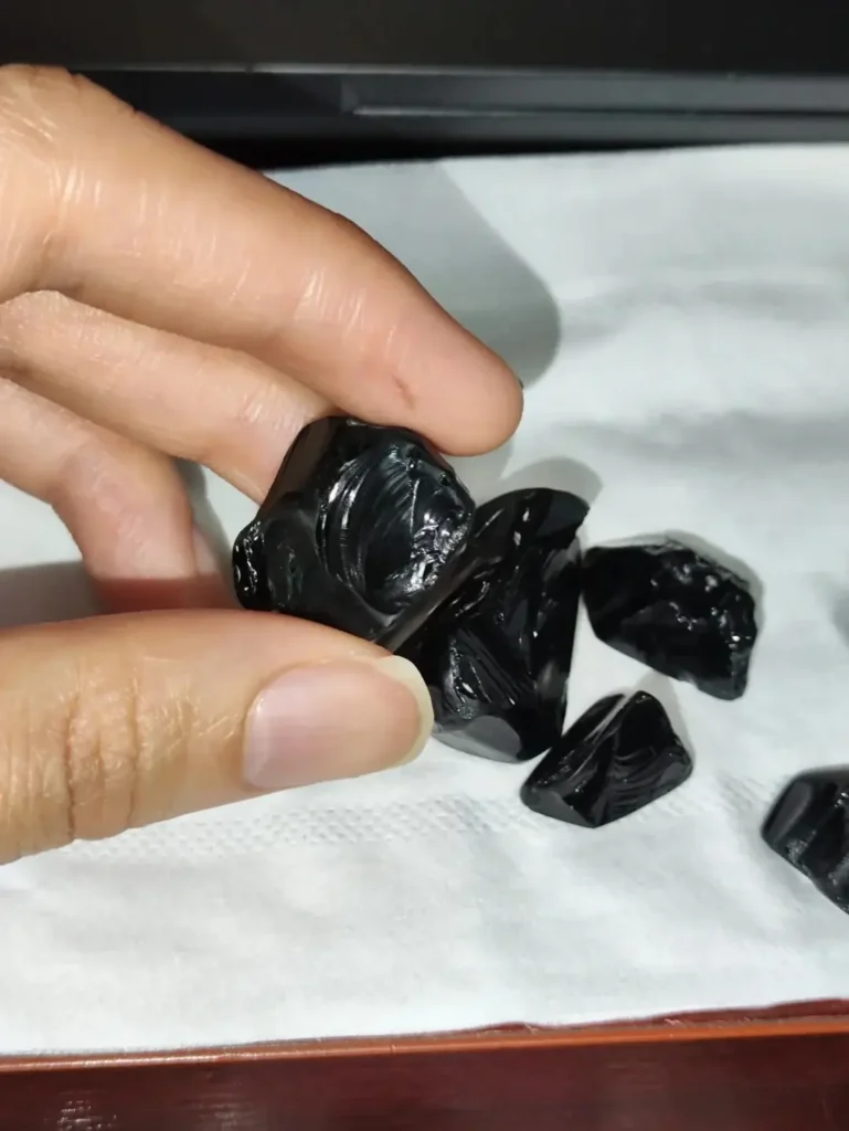A hand holding a piece of raw black obsidian showing shiny conchoidal fractures.